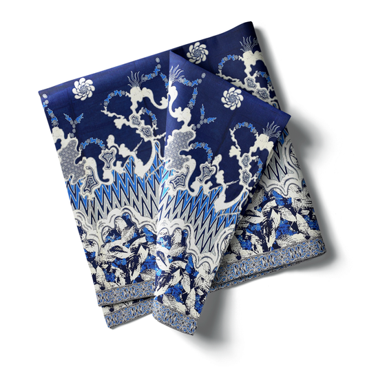 kain Katun Zig-Zag Daun dengan motif abstrak bergelombang, masing-masing dengan kombinasi warna yang berbeda (biru, cokelat, dan hijau). Kain dilipat dan diletakkan di samping hiasan pinus